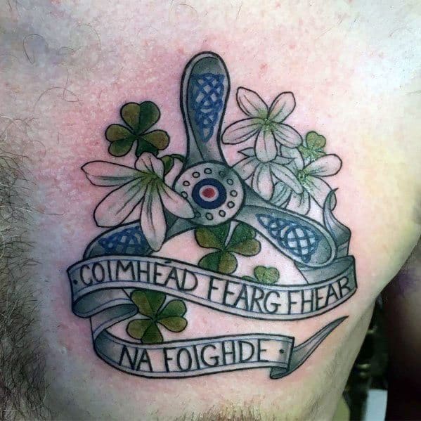 Colorful chest tattoo featuring a Celtic-style propeller, shamrocks, white flowers, and a Gaelic banner reading "Coimhead Fearg Fhear na Foighde."