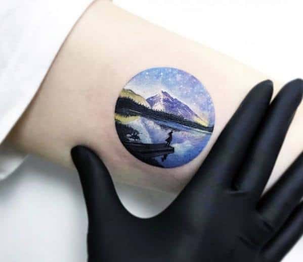 Small circular tattoo of a person fishing on a dock, with a starry sky, calm lake, and snow-capped mountains reflected in the water.