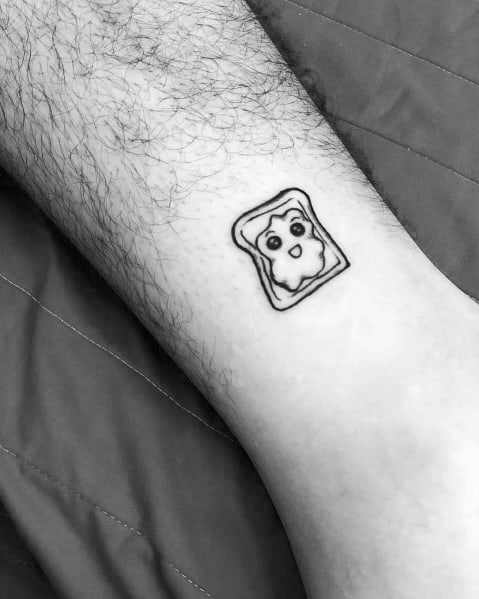 Small minimalist toast tattoo with a playful face in the butter, outlined in black for a simple yet quirky design on the lower leg.