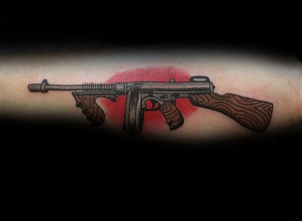 Traditional-style Tommy gun tattoo with bold black outlines, wood grain details, and a red background for a striking contrast on the forearm.