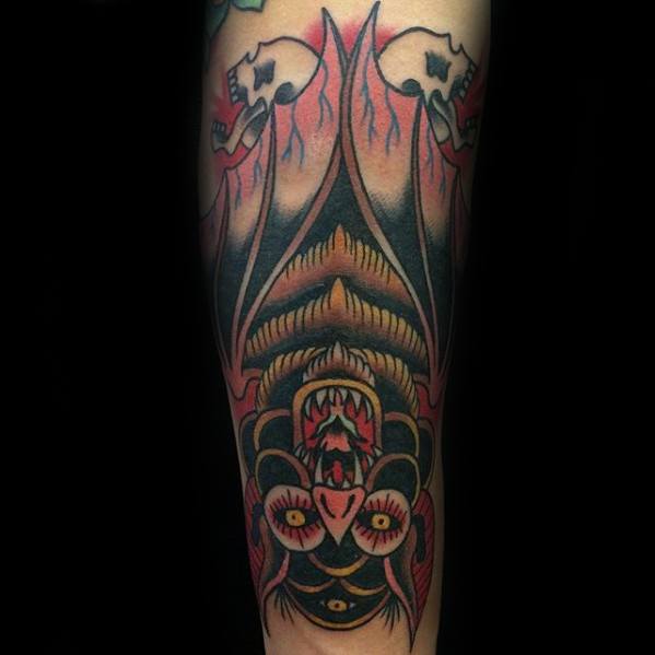 Traditional upside-down bat tattoo with fiery background, red and yellow tones, flanked by skulls and bold symmetrical wings.