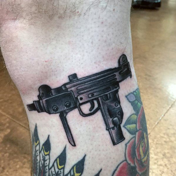 Realistic black and grey Uzi tattoo on the leg with bold shading and fine details, blending seamlessly with surrounding colorful ink.