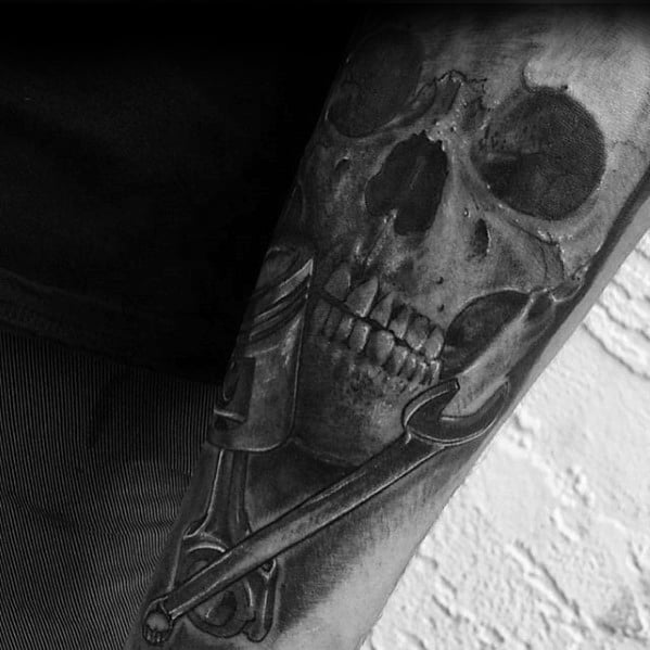 Realistic black and grey tattoo of a skull partially merged with a wrench on the forearm, creating a dark, mechanical fusion design.