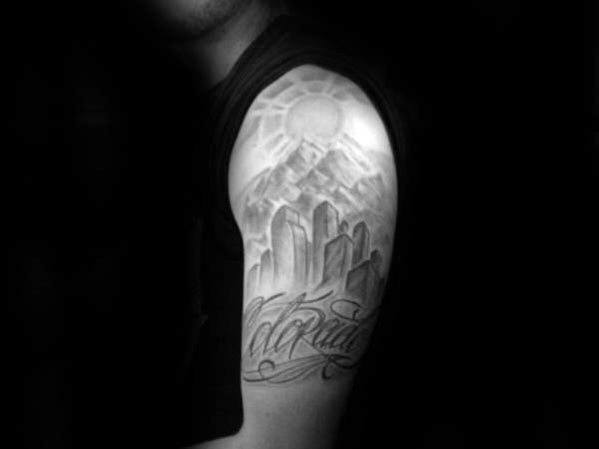 Black and grey Denver skyline tattoo on upper arm with mountains, a radiant sun, and elegant “Colorado” script below.