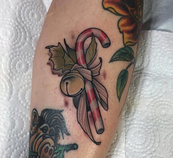 Christmas tattoo of a candy cane tied with a ribbon, featuring holly leaves and a jingle bell, blending festive holiday charm with a classic design.
