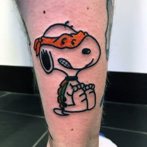 Colorful tattoo of Snoopy dressed as a Ninja Turtle, wearing an orange mask and holding a nunchuck, placed on a person's leg.