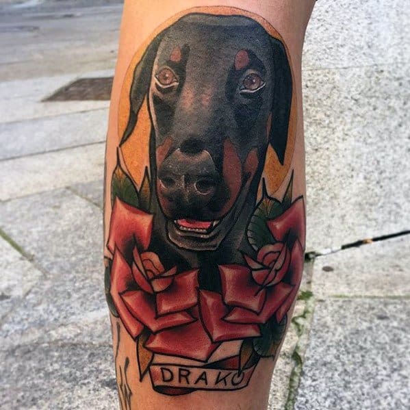 Traditional Doberman tattoo with bold black and red shading, surrounded by roses and a name banner reading “Drako” for a heartfelt tribute.