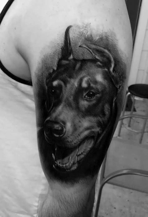 Realistic black and grey Doberman tattoo on the upper arm, showing a joyful expression with smooth shading and lifelike detail.