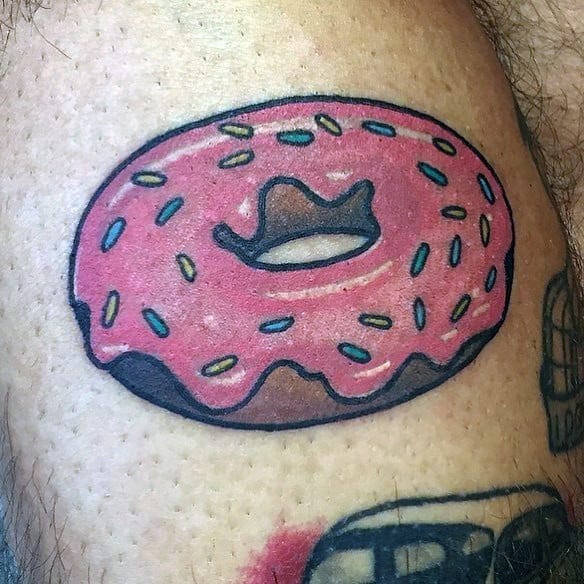 Tattoo of a simple pink frosted donut with sprinkles and bold black outlines, done in a playful cartoon style.