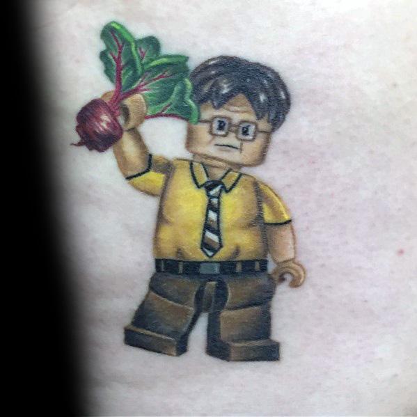 Tattoo of Dwight Schrute as a LEGO figure, wearing a yellow shirt and tie, holding a beet in one hand, with detailed shading and bold colors.