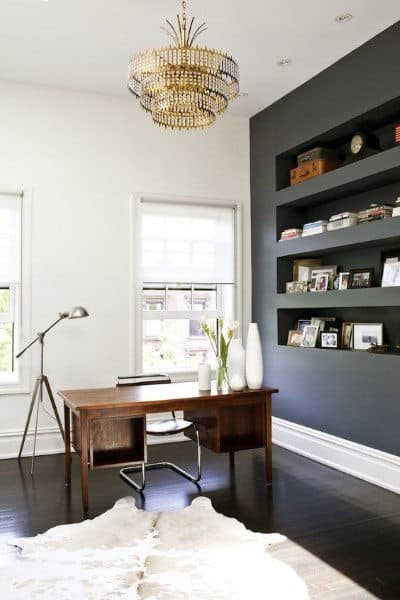 Modern home office with dark wood desk, white chair, chandelier, dark accent wall with shelves, and cowhide rug