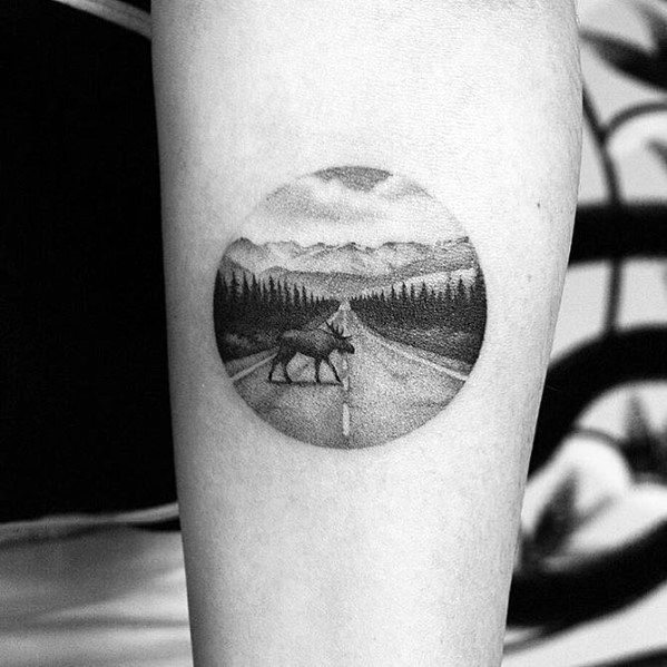 Minimalist black and grey elk tattoo in a circular frame, featuring a detailed mountain road scene with fine shading for a serene and artistic design.