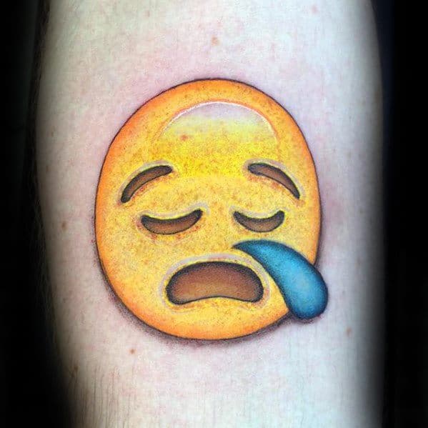 Tattoo of a tired emoji with closed eyes, frowning mouth, and a blue droplet on a person's arm