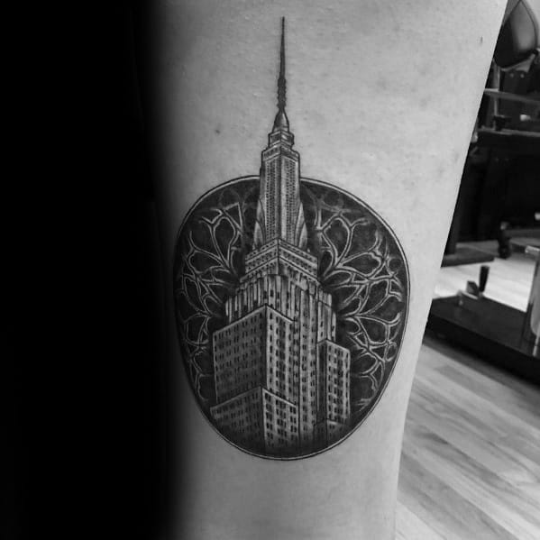 Empire State Building tattoo enclosed in a detailed circular frame with intricate patterns, inked in bold black shading.