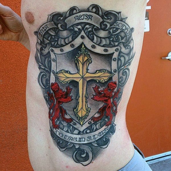 Side torso tattoo of a medieval-style shield with a golden cross, two red lions, ornate scrollwork, and banners reading names and “Nemo Malum Superat.”