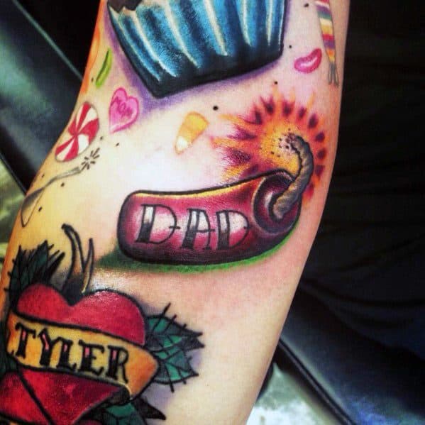 Tattoo of a lit firecracker with "DAD" written on it, surrounded by colorful elements like a cupcake and candy, creating a bold, playful tribute.
