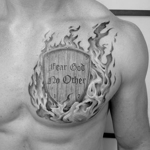 Chest tattoo of a wooden shield surrounded by flames, engraved with the words “Fear God No Other” in a bold, gothic-style font.