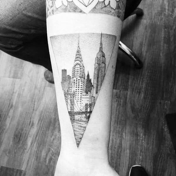 Forearm tattoo of the New York City skyline inside a triangle, featuring the Empire State and Chrysler buildings in fine black ink.