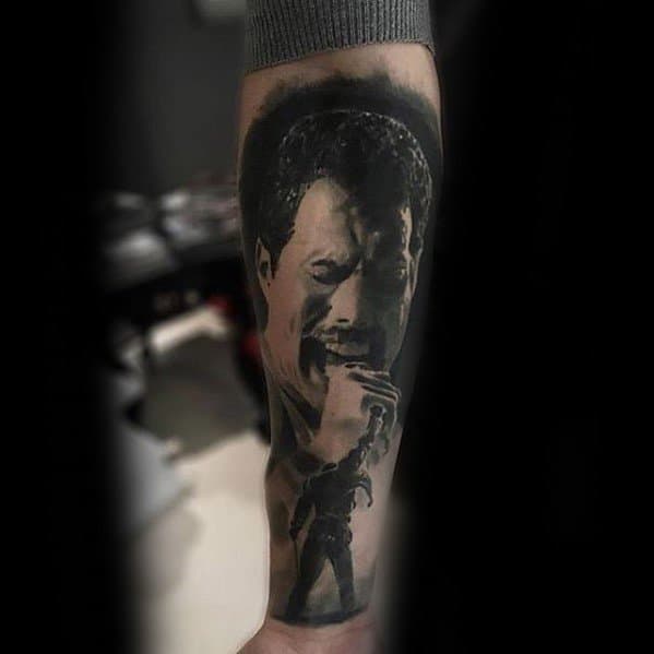 Black and grey forearm tattoo of Freddie Mercury with a powerful facial expression, paired with a smaller full-body stage pose below.