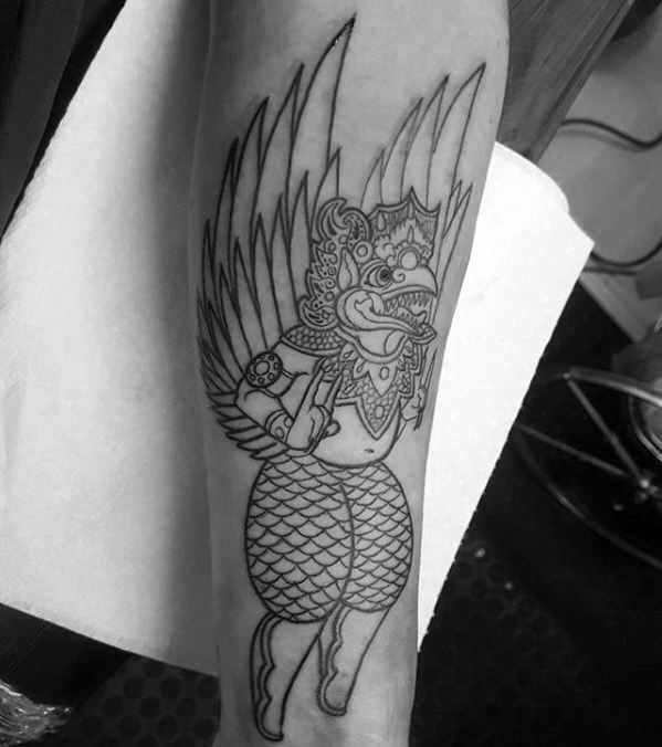 Garuda tattoo outline on the forearm featuring a playful winged figure with traditional attire, detailed headgear, and expressive face.
