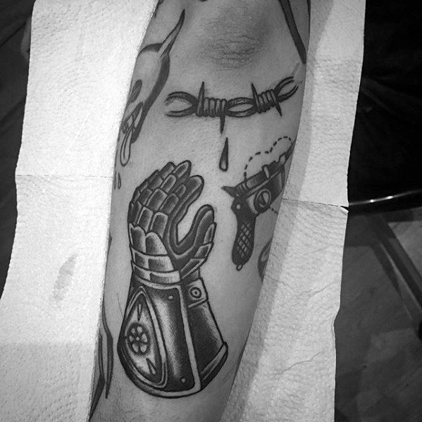 Black ink tattoo of a robotic-style gauntlet in a praying position, placed near barbed wire and gun designs, symbolizing contrast.