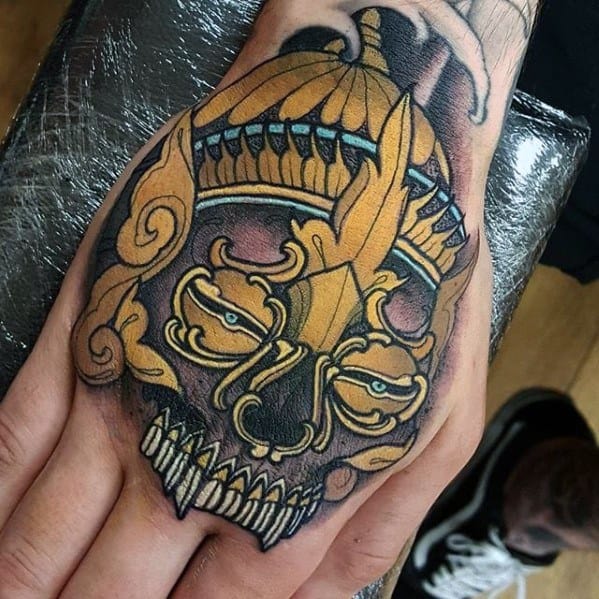 Ornate Tibetan skull tattoo on the hand with golden crown, sharp teeth, and bold black outlines for a striking, intense look.