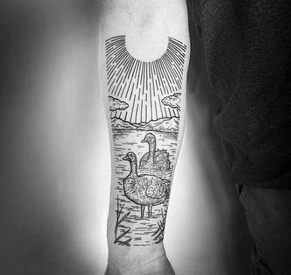 Black and gray tattoo of two geese in a lake with mountains, clouds, and a radiant sun, inked on the forearm in a fine line illustrative style.