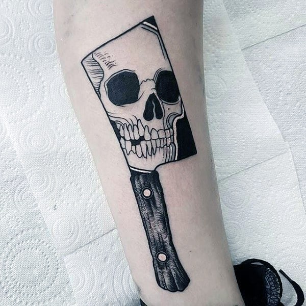 Black and grey tattoo featuring a cleaver with a skull reflection in the blade, combining horror and gothic elements with bold linework.
