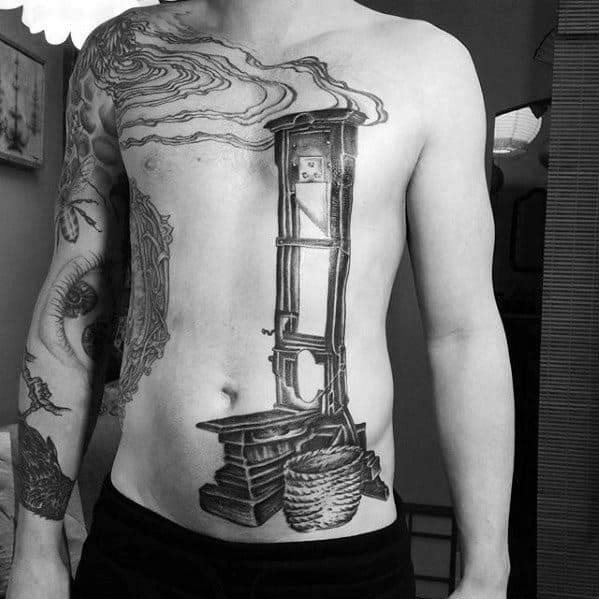 Massive black and grey guillotine tattoo spanning the torso, featuring stacked books, a detailed basket, and swirling smoke above the blade.