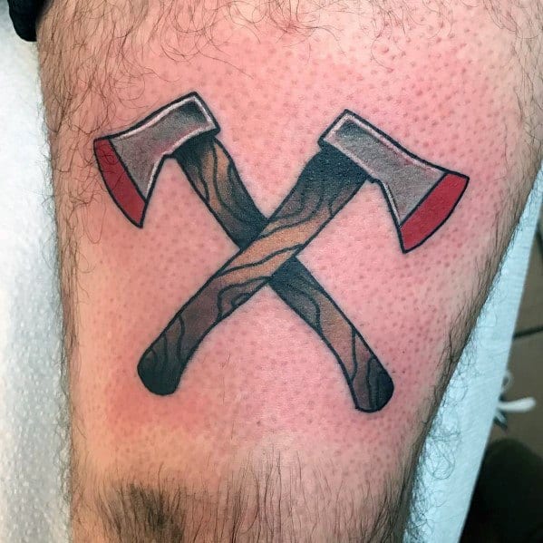 Traditional-style tattoo of two crossed hatchets with wood-textured handles and red-tipped blades, inked with bold outlines and shading on the thigh.