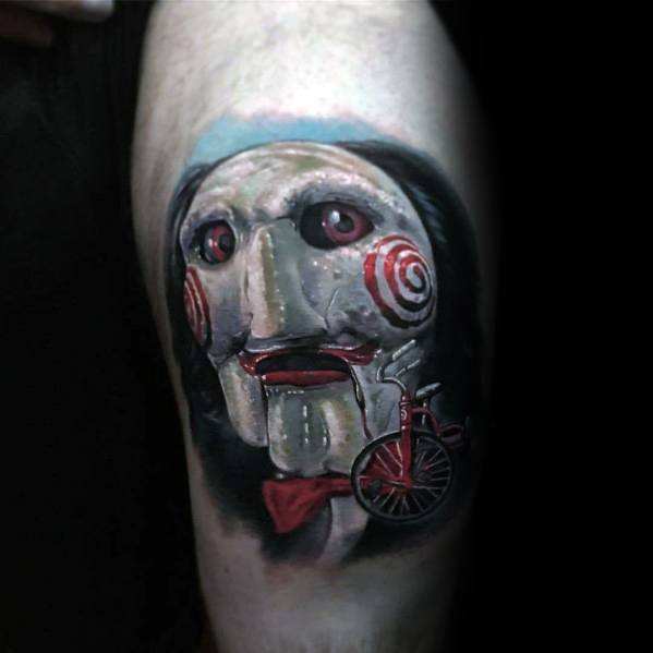 Realistic color tattoo of Billy the Puppet from *Saw*, featuring his eerie red spiral cheeks, glowing eyes, and signature tricycle for a chilling horror design.