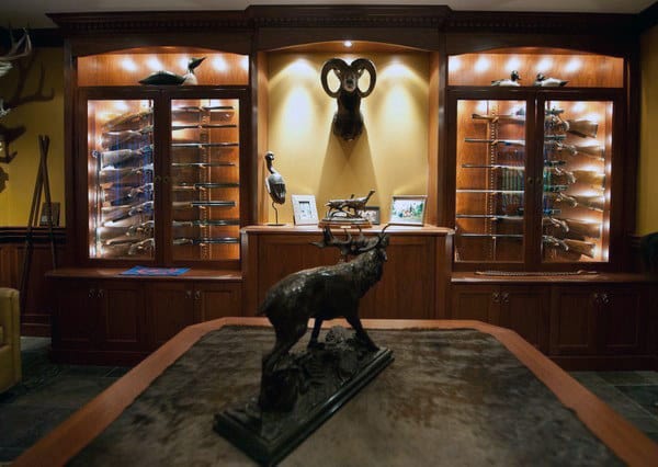 A hunting-themed gun room featuring illuminated wood cabinets, wildlife decor, and a centerpiece table with a deer sculpture.