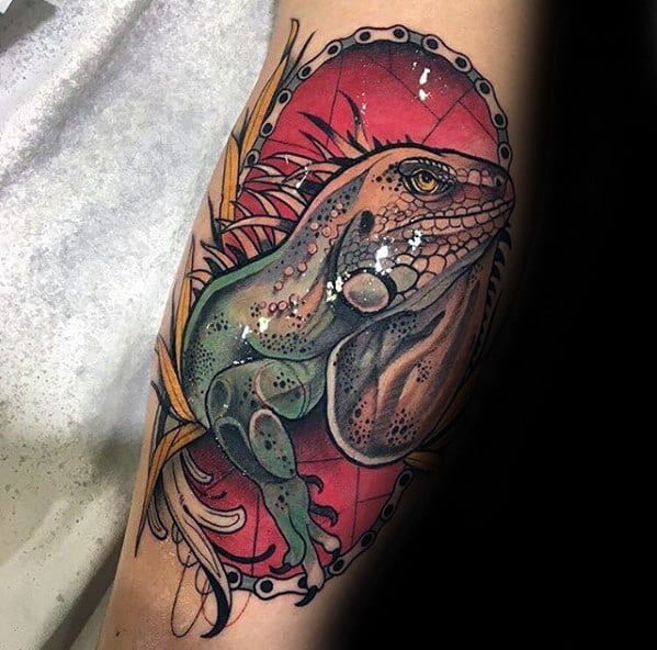 Neo-traditional iguana tattoo with green and beige tones, framed in an ornate oval border over a bold red background with leaf accents.