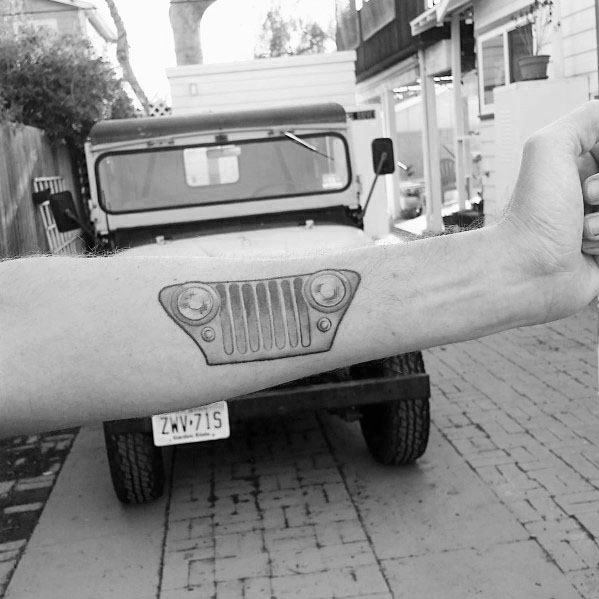 Realistic black and grey tattoo of a Jeep grille on the forearm, positioned to align with an actual Jeep in the background for a creative effect.