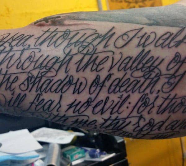 Large cursive script tattoo on the inner bicep featuring a passage from Psalm 23:4, 'Though I walk through the valley of the shadow of death