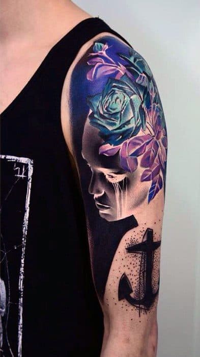 Tattoo of a pensive face in black and white with blue and purple roses blooming from the head, paired with a bold black anchor.