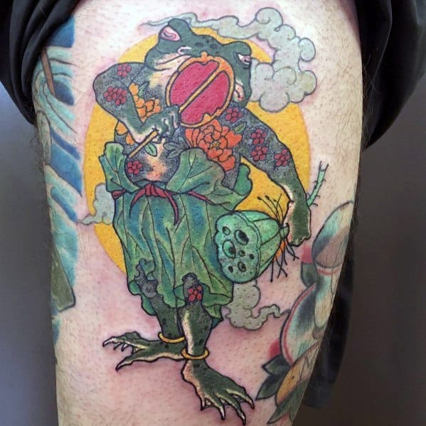 Japanese-style tattoo featuring a warrior frog dressed in traditional robes, holding a red fan and a lotus pod, with a golden halo and swirling clouds.