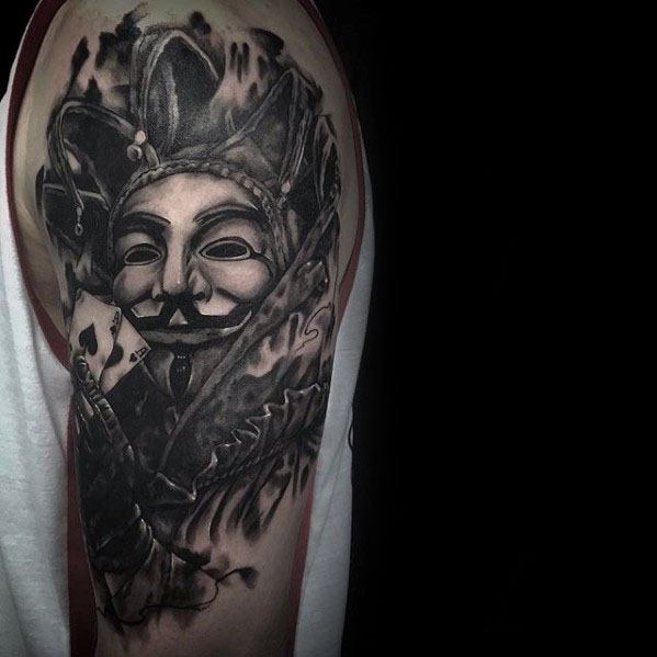 A black and gray tattoo of a jester wearing a mask, holding a playing card, with flowing details and intricate shading, placed on the upper arm.