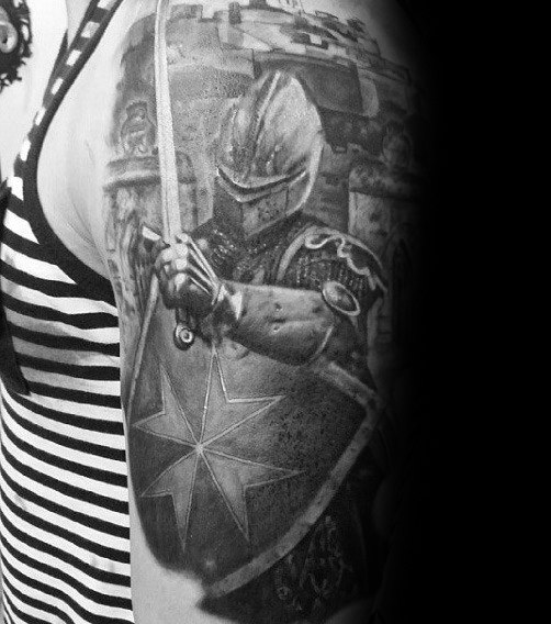 Realistic black and grey upper arm tattoo of a medieval knight in full armor holding a sword and a shield with a prominent Maltese cross.