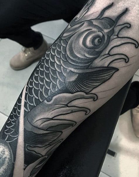 A black and grey tattoo featuring a detailed koi fish with intricate scales, swimming amidst stylized waves, located on the forearm