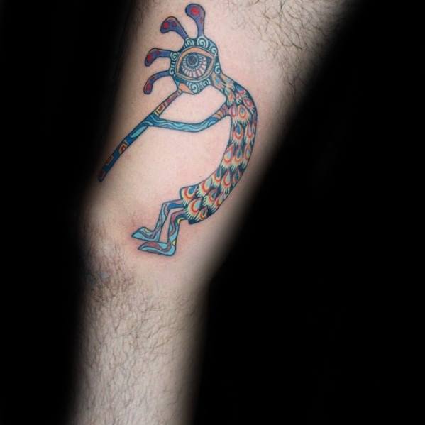 Colorful Kokopelli tattoo featuring a tribal design with vibrant feathers, an intricate eye detail on the head, and geometric accents on the body.