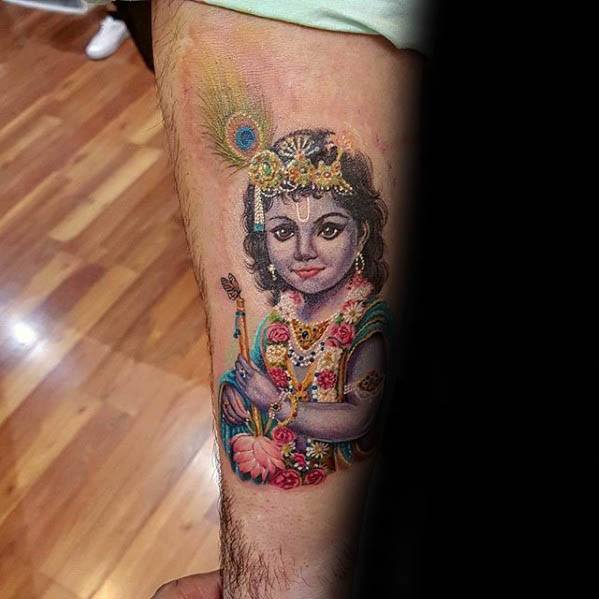 Colorful tattoo of young Krishna holding a flute, adorned with floral garlands, a peacock feather, and a gentle smile on the forearm.