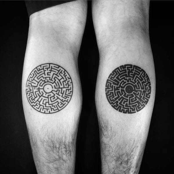 Matching circular labyrinth tattoos on a man's calves, featuring intricate maze designs with one in negative space and the other filled with black ink.