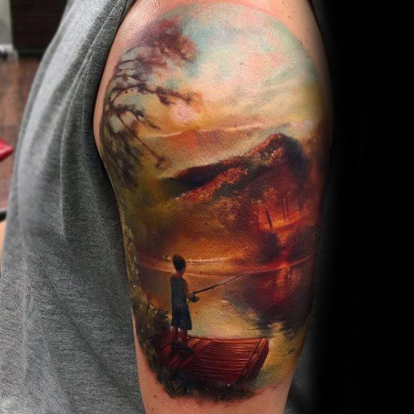 Realistic upper arm tattoo of a child fishing from a dock at sunset, with warm hues, misty trees, and mountains reflected in a calm lake.
