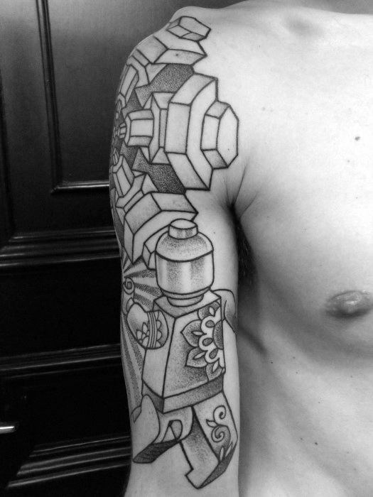 Black linework tattoo wrapping from shoulder to upper arm, featuring a Lego minifigure with floral patterns and abstract geometric Lego shapes above.