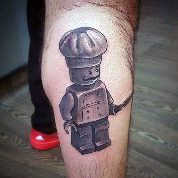 Black and grey tattoo of a Lego chef minifigure holding a knife, featuring detailed shading on the hat, jacket, and limbs for a 3D look.