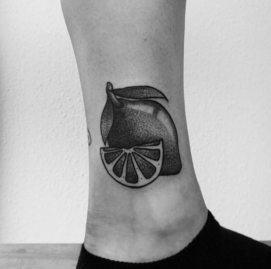 Black and grey ankle tattoo of a lemon and a lemon slice with bold outlines and smooth dotwork shading for a clean, minimal look.