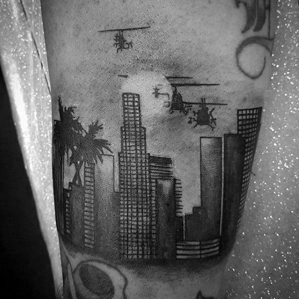 Black and gray tattoo of Los Angeles skyline with palm trees, flying helicopters, full moon, and a dramatic night sky.