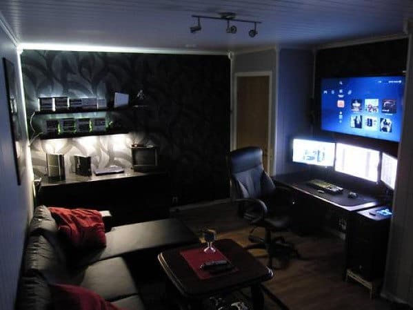 A cozy gaming room with dim lighting, a triple-monitor setup, black couch, and wall-mounted TV