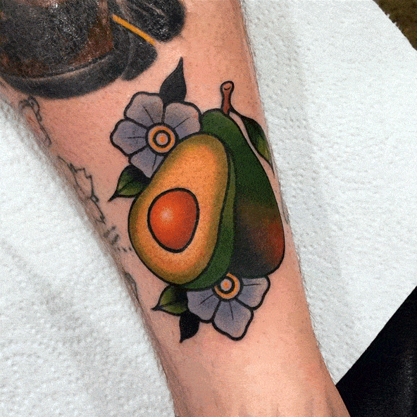 Bold traditional avocado tattoo on the forearm with vibrant green, orange, and red tones, accented by white flowers and deep shading.